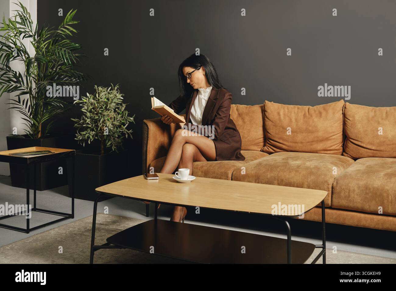 Eine Frau in einem schicken Outfit genießt einen ruhigen Moment mit einem Buch auf einer gemütlichen Couch. Das moderne Wohnzimmer ist mit Grün und minimalistischem Pelz ausgestattet Stockfoto