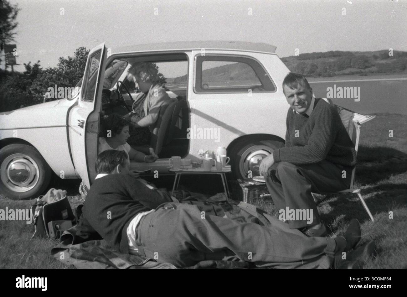 1960er Jahre, historisch, auf grasbewachsenem Land am See, eine Familie, die ein Picknick vor ihrem Auto aus der damaligen Zeit machte, ein Austin A40 Farina, hergestellt von der British Motor Corporation (BMC). Der dreitürige Kleinwagen wurde von 1958 bis 1967 produziert. Stockfoto