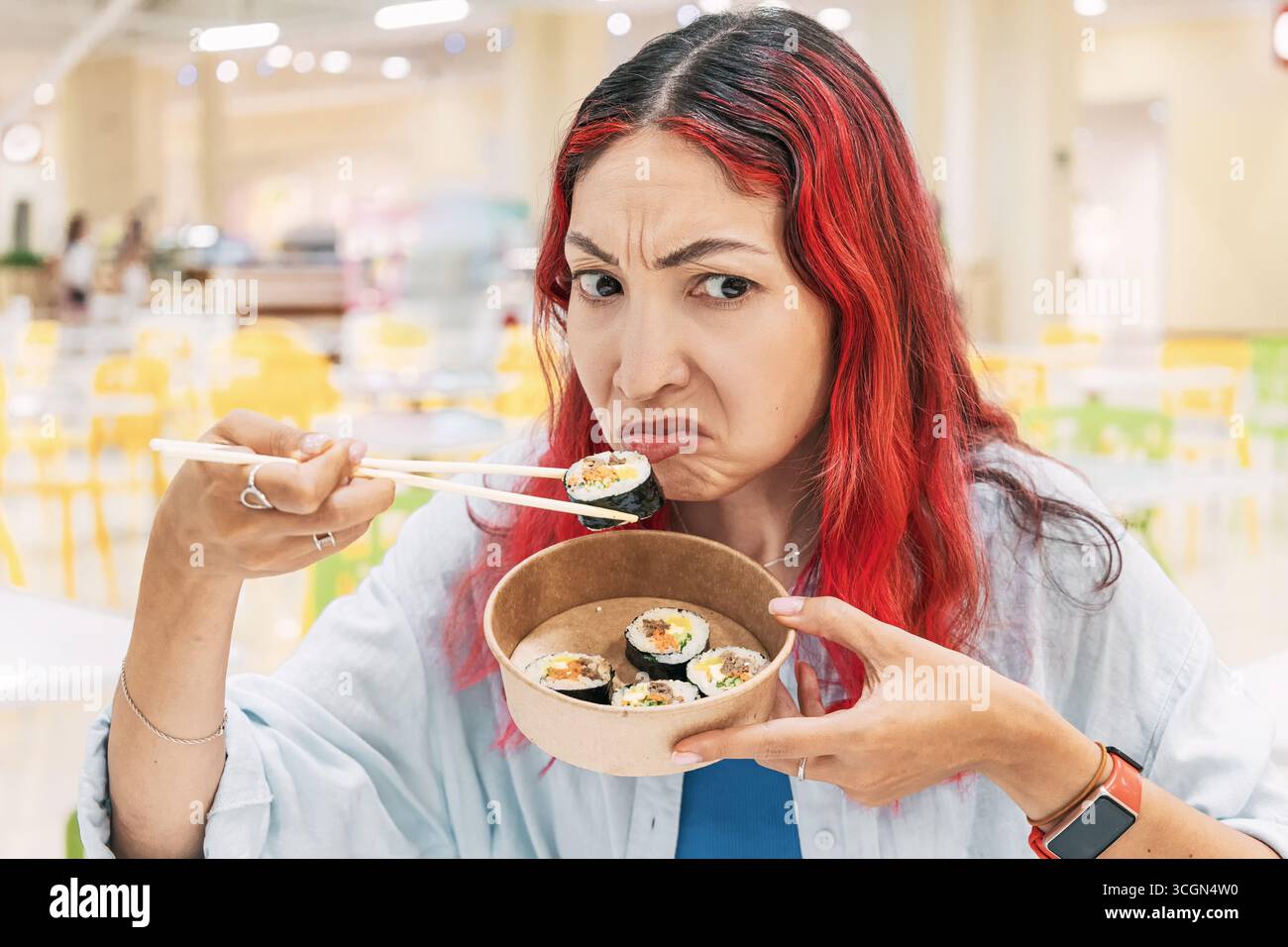 Eine junge Frau, die in einem belebten Speisesaal Kimbap oder Sushi mit Essstäbchen isst und ihre Unzufriedenheit über verdorbenes Essen zum Ausdruck bringt Stockfoto