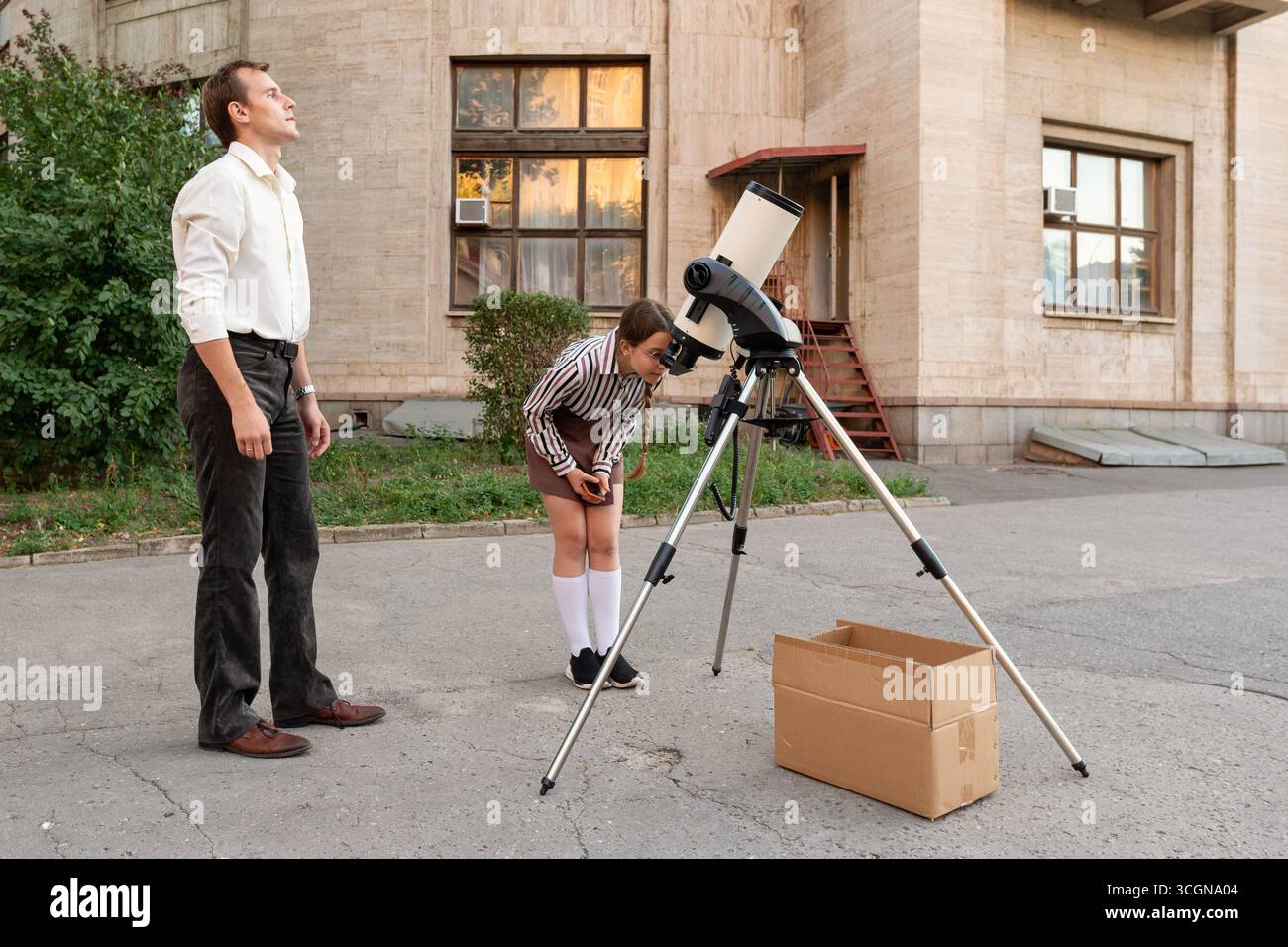 Während eines Astronomiekurses außerhalb eines Gebäudes beobachtet eine Schülerin sorgfältig die Teleskopeinstellungen unter Anleitung ihres Lehrers und konzentriert sich dabei auf Präzision für Stockfoto