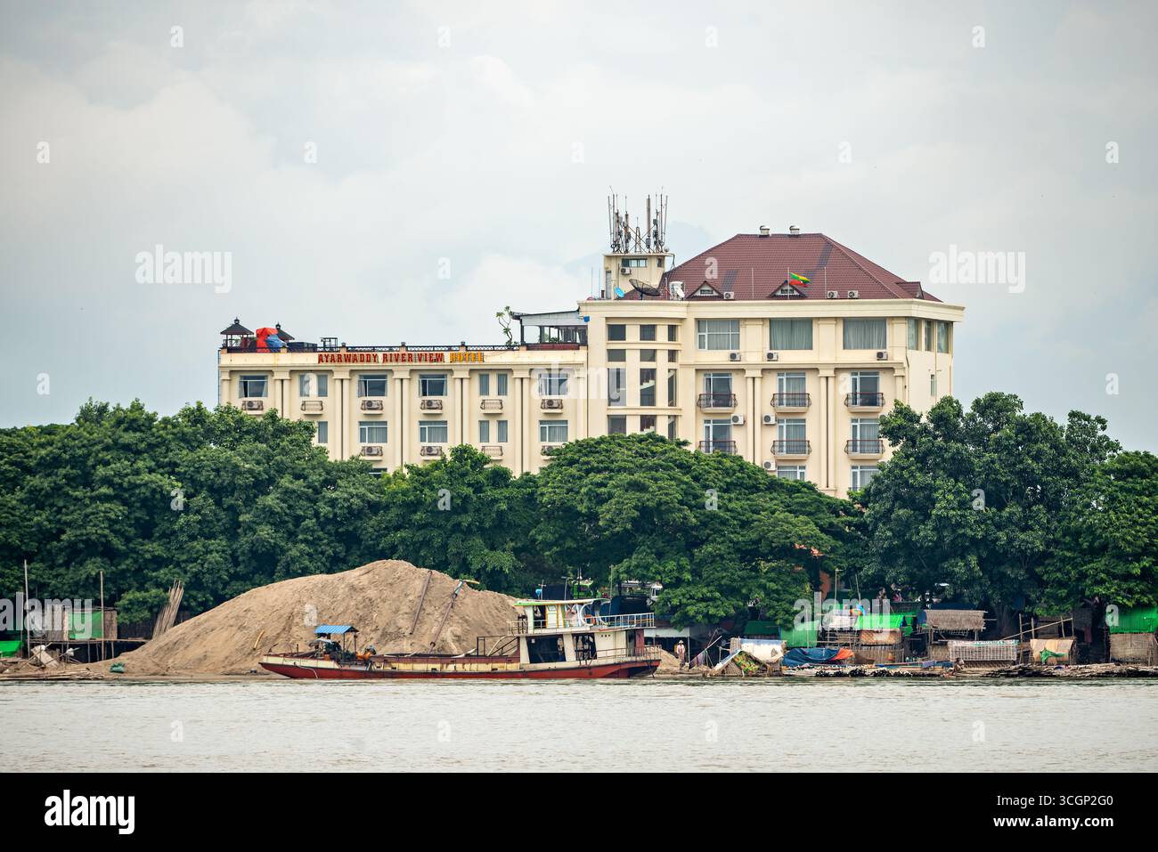 Ayeyarwaddy River View Hotel and Sand Barge on Ayeyarwaddy River Mandalay Myanmar // MANDALAY, Myanmar – das Ayarwaddy Riverview Hotel (auch bekannt als Ayeyarwaddy River View Hotel) ist neben einem Sandkahn auf dem Ayeyarwady River (auch bekannt als Irrawaddy River) in Mandalay zu sehen. Das Hotel bietet Unterkünfte mit Blick auf Myanmars wichtigste kommerzielle Wasserstraße. Sandkähne wie dieser werden häufig für den Transport von Baumaterial entlang des Flusses verwendet. Der Ayeyarwady River ist eine wichtige Verkehrs- und Wirtschaftsstraße und fließt etwa 2.170 Kilometer (1.350 Meilen) durch Stockfoto