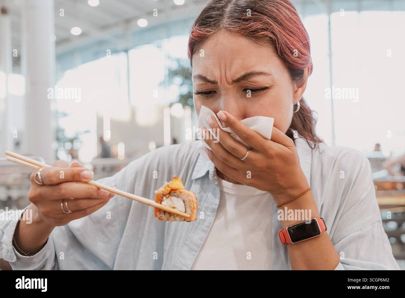 Die junge Frau hält eine Sushi-Rolle mit Essstäbchen und bedeckt ihre Nase mit einer Serviette und drückt Ekel aus Stockfoto