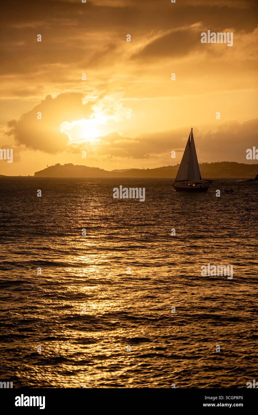 Segelboot bei Sonnenuntergang über Wasser in der Nähe von St. Thomas von Cruz Bay St. John USVI // St. JOHN, USVI — Ein Segelboot navigiert durch die Gewässer vor St. John, U.S. Virgin Islands, während die Sonne untergeht und ein goldenes Leuchten über das Karibische Meer wirft. In der Ferne ist die Insel St. Thomas am Horizont sichtbar. Die Aussicht wird vom Galgen Point in Cruz Bay erfasst, einem beliebten Aussichtspunkt auf St. John, der für seine malerischen Ausblicke bekannt ist. Die ruhige Szene zeigt die natürliche Schönheit der U.S. Virgin Islands, einem Gebiet mit mehr als 50 Inseln und Inseln. Dieser Archipel liegt im Nordosten der Karibik Stockfoto