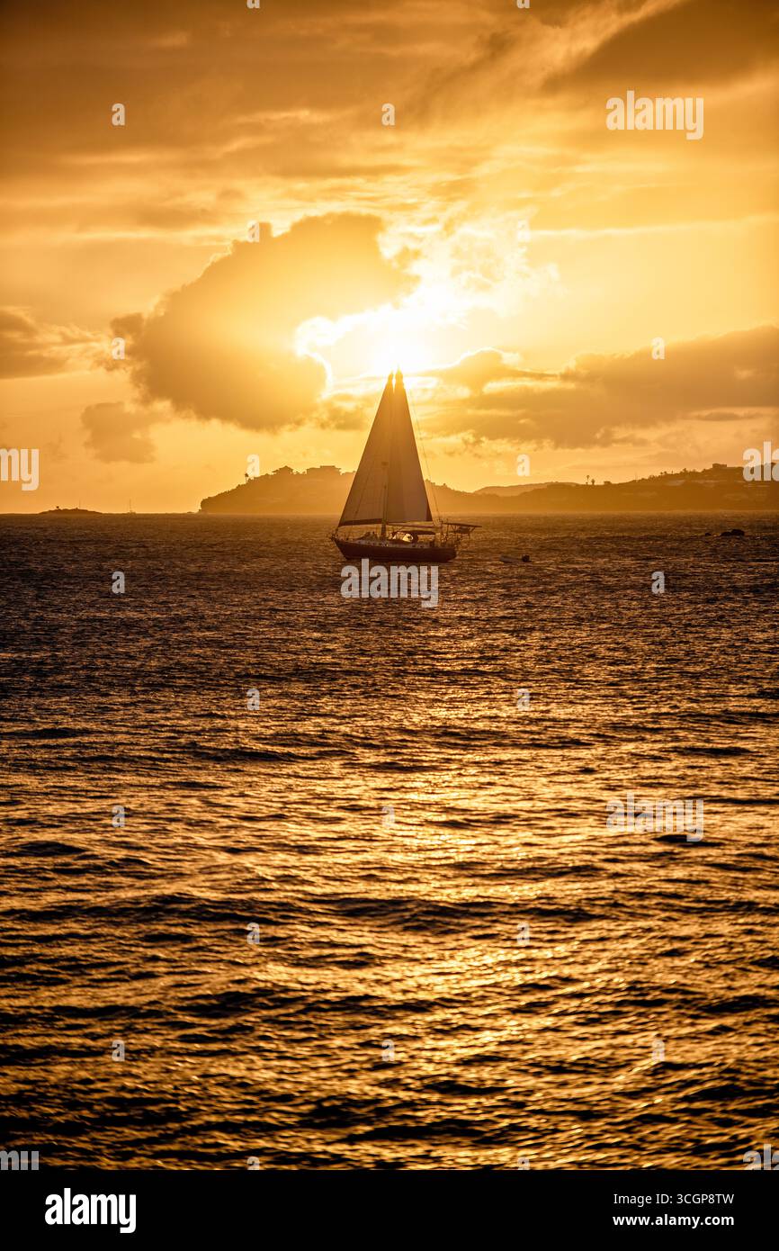 Segelboot bei Sonnenuntergang über der Karibik St. John USVI // St. JOHN, USVI — Ein Segelboot fährt bei Sonnenuntergang durch die Karibik, wobei die Insel St. Thomas in der Ferne sichtbar ist. Die Aussicht ist vom Galgen Point in Cruz Bay auf St. John. Die Sonnenstrahlen brechen durch die Wolken und werfen ein warmes, goldenes Licht über das Wasser. St. John ist eine der Hauptinseln der United States Virgin Islands, einem Territorium der Vereinigten Staaten auf den Kleinen Antillen. Die Inseln sind bekannt für ihre natürliche Schönheit und ein beliebtes Ziel für Segeln und Wassersport. Stockfoto