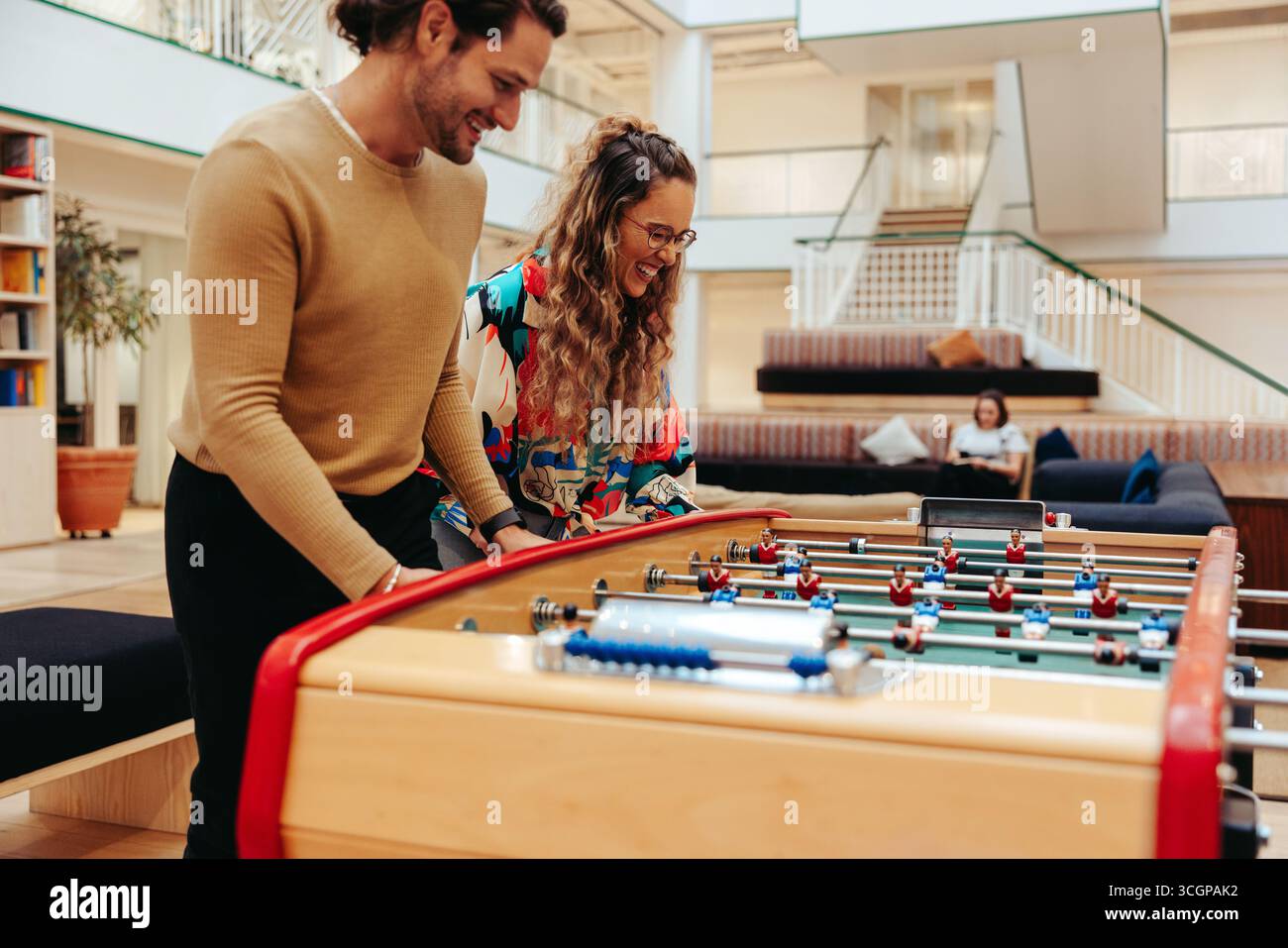 Zwei Kollegen teilen sich während der Mittagspause einen Moment des Spaßes und der Entspannung, indem sie in einem hellen, modernen Büro Tischfußball spielen. Dieses Bild zeigt das Jo Stockfoto