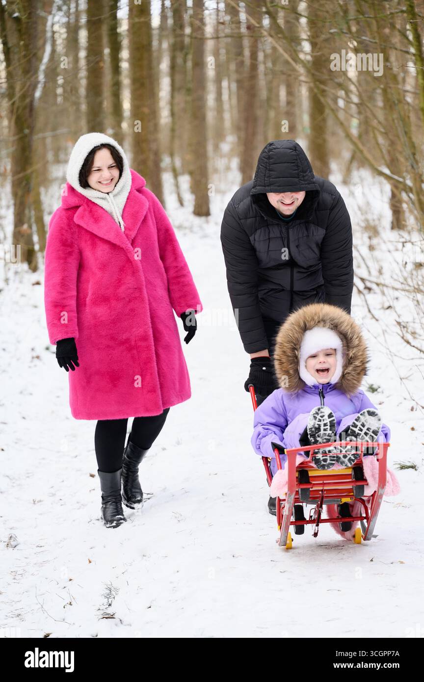 Familienspaziergang durch den verschneiten Park. Vater schiebt Mädchen, die auf Kinderschlitten sitzen, Mutter läuft neben ihr und lacht glücklich. Winterspaß im Freien, Weihnachtsurlaub mit der Familie Stockfoto