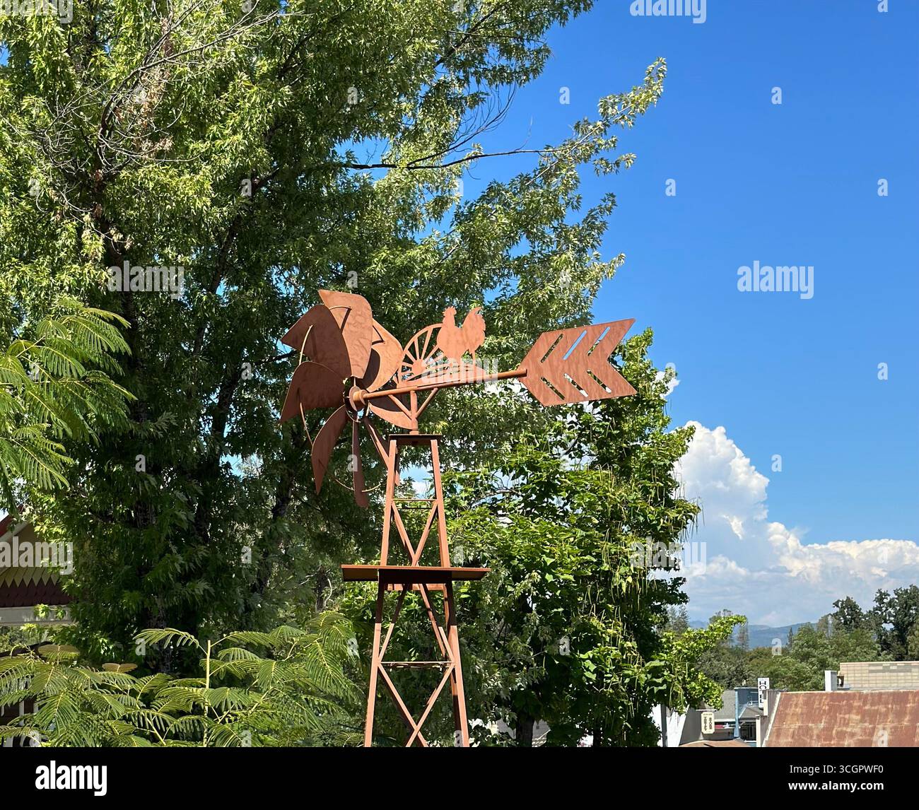 Eine rustikale Windmühle aus Metall mit Hahnendesign, fotografiert im Freien vor einem hellblauen Himmel und grünen Bäumen. - Smartphone-aufgenommenes Stockfoto