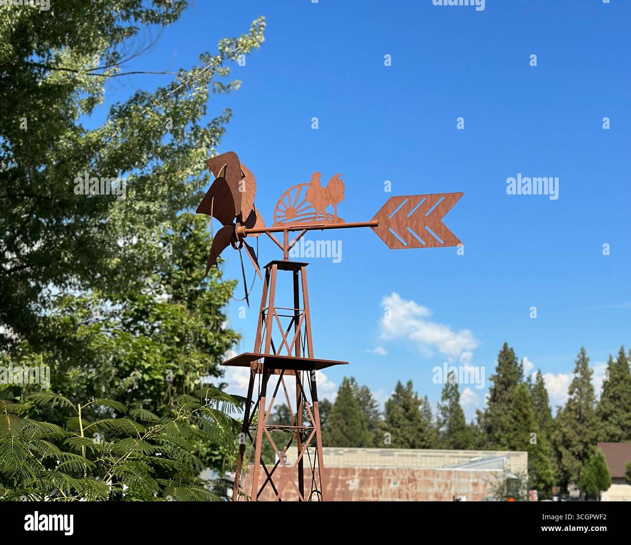 Eine rustikale Windmühle aus Metall mit Hahnendesign, fotografiert im Freien vor einem hellblauen Himmel und grünen Bäumen. - Smartphone-aufgenommenes Stockfoto