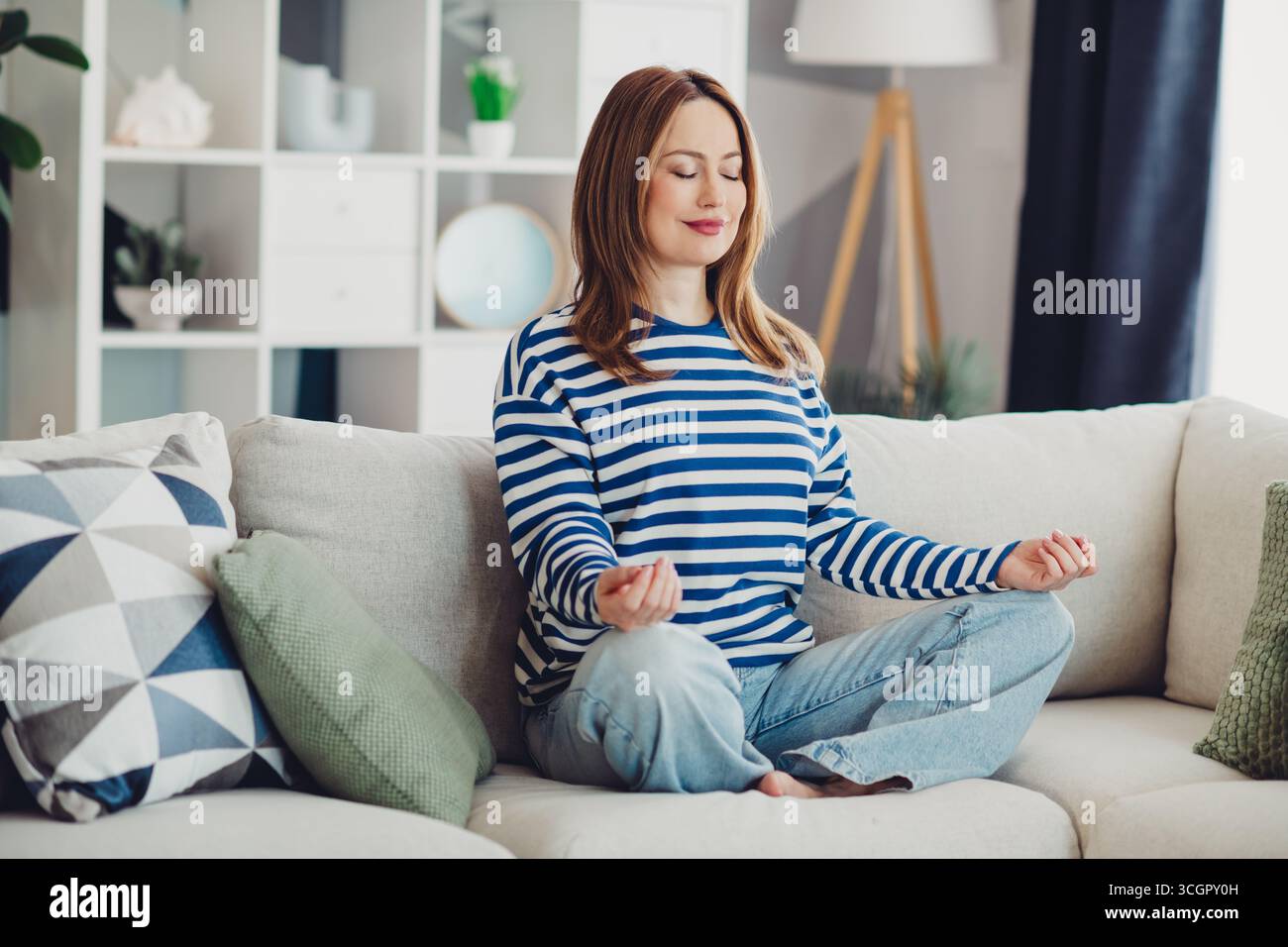 Junge Frau übt Entspannungstechniken auf einer gemütlichen Couch zu Hause in einem wunderschön eingerichteten Wohnzimmer Stockfoto
