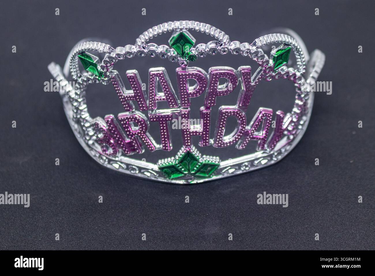 Metallische Happy Birthday Crown mit rosa Text und grünen Edelsteinen auf schwarzem Hintergrund Stockfoto