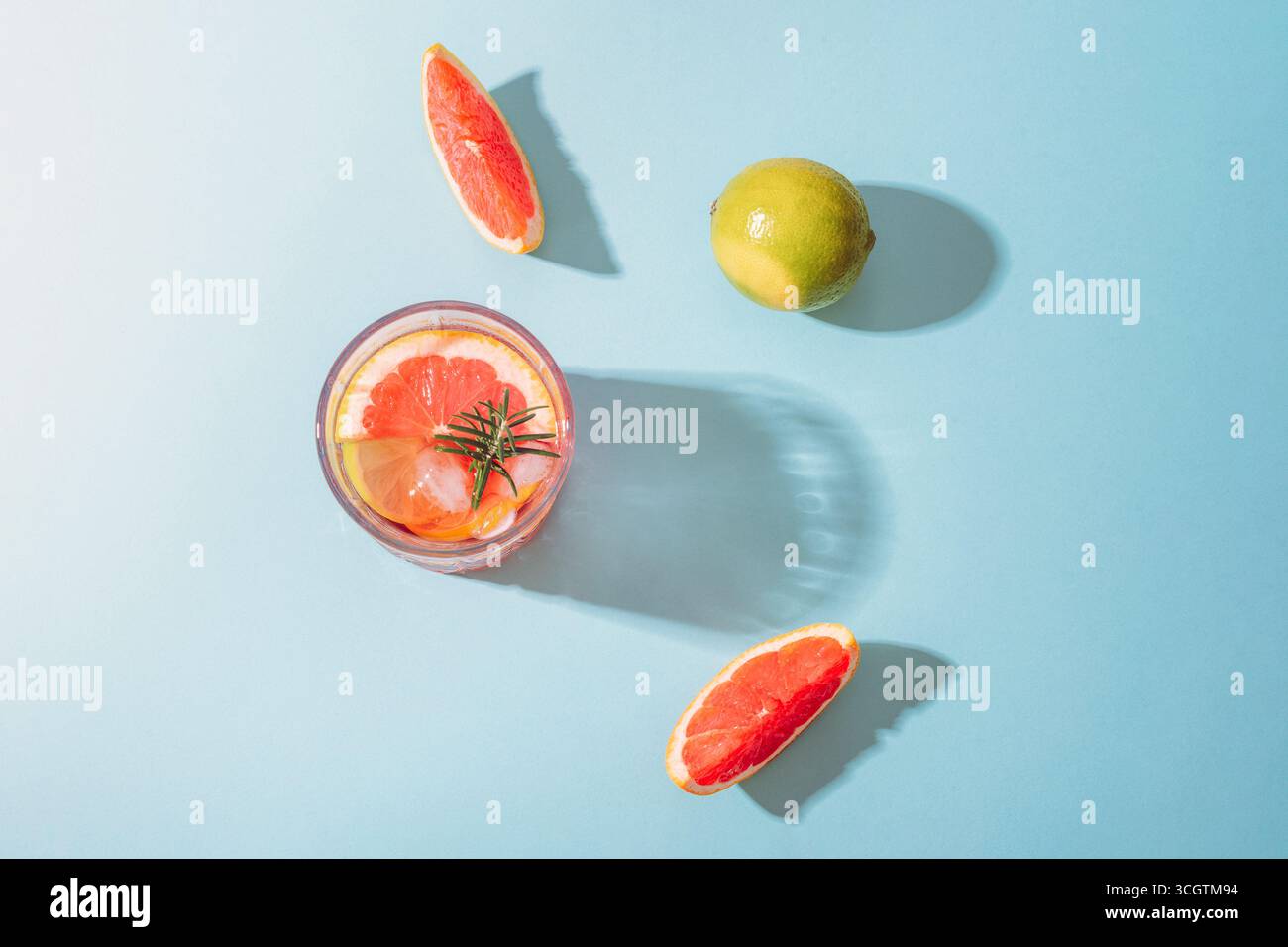 Glas Grapefruitcocktail mit Limettenscheiben und Rosmarin auf blauem Hintergrund mit scharfen Schatten. Draufsicht, flach. Stockfoto