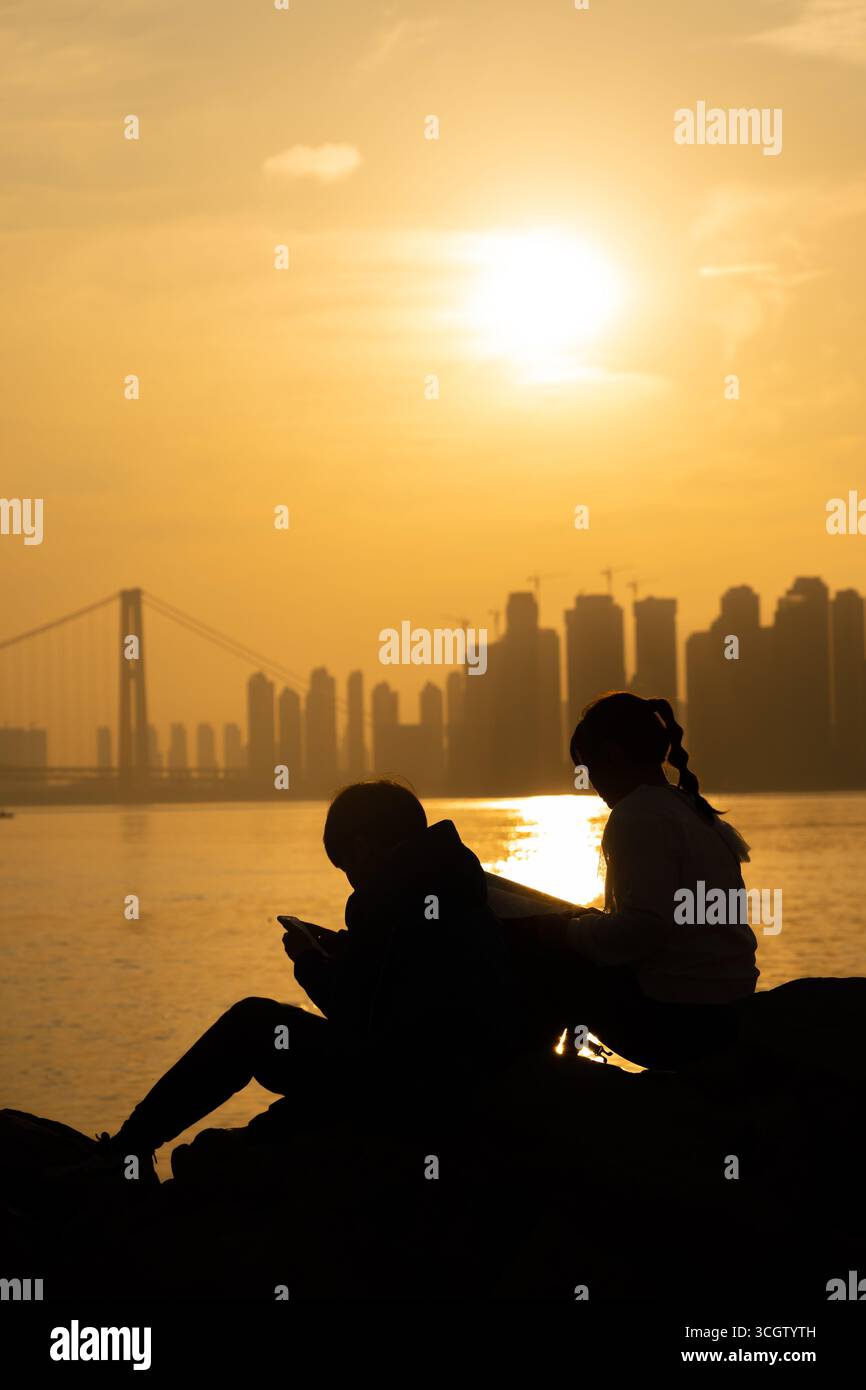 Zwei Figuren stehen vor einem leuchtend orangefarbenen Sonnenuntergang, die auf Felsen in der Nähe des Wassers sitzen. Die Szene vermittelt ein Gefühl der Ruhe und des gemeinsamen Erlebnisses Stockfoto