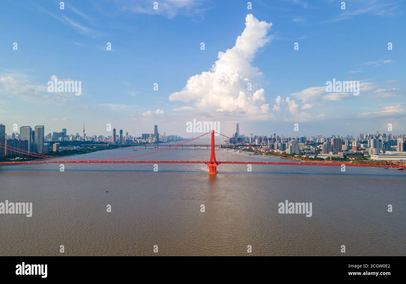 Die Skyline der Stadt Wuhan erstreckt sich über den Yangtze-Fluss in China und bietet eine leuchtend rote Hängebrücke mit hohen Türmen vor blauem Himmel und Fluf Stockfoto