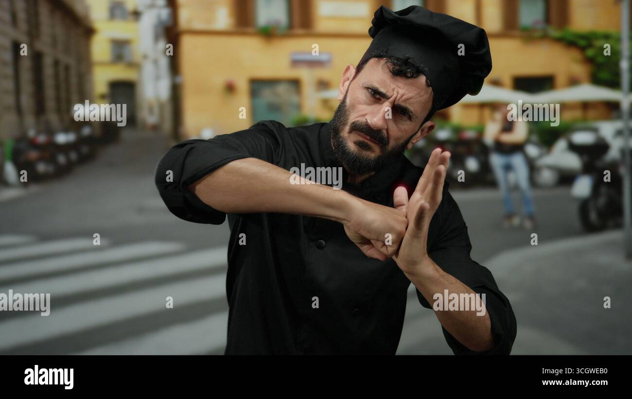 Bärtiger hispanischer Mann, der als Koch gekleidet ist und Boxpositionen auf der Straße im Freien nachahmt und professionelle Kleidung mit spielerischem Ausdruck verbindet. Stockfoto