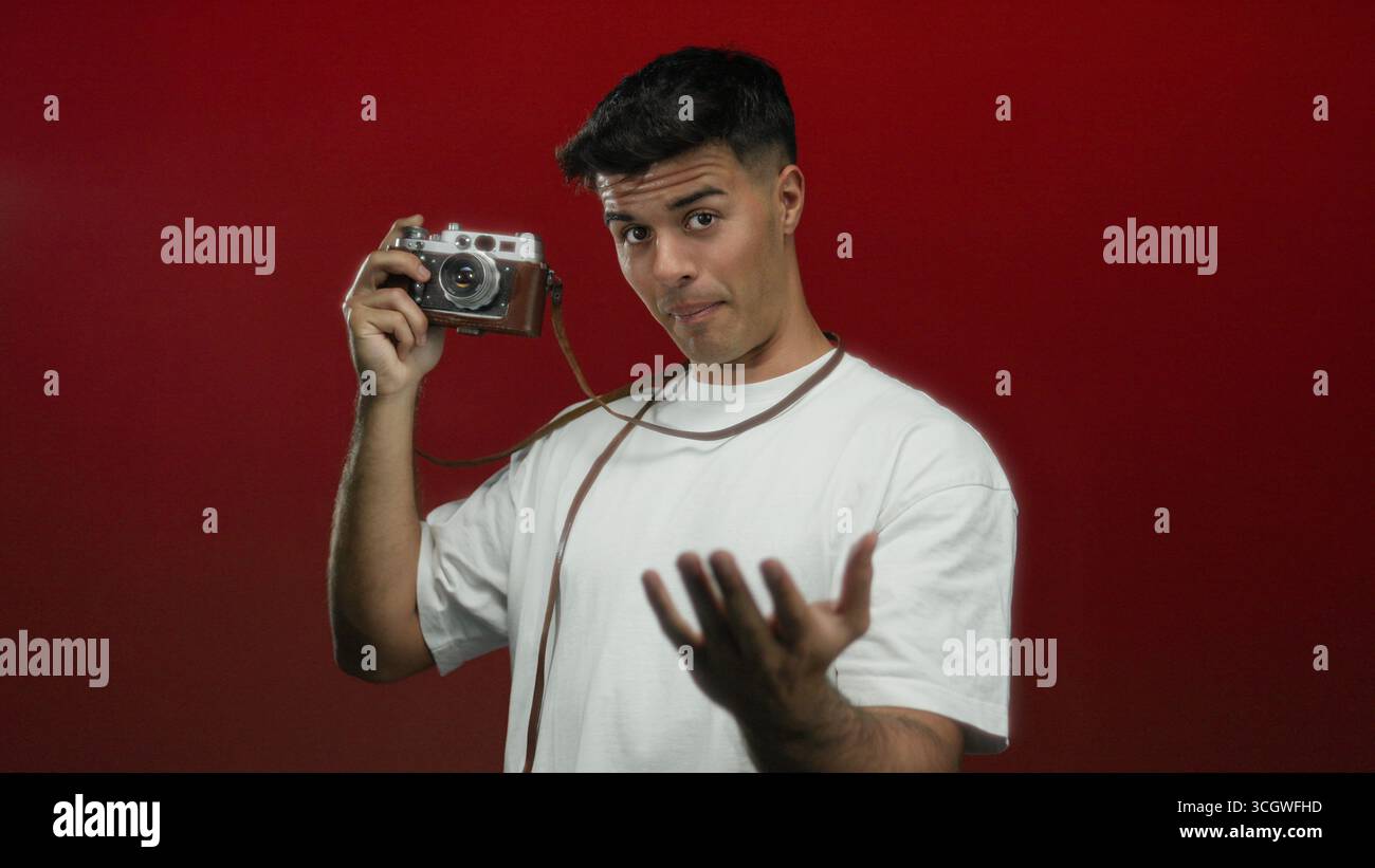 Junger Mann mit einer alten Kamera, der eine Geste über isoliertem rotem Hintergrund macht und einen selbstbewussten und einladenden Ausdruck zeigt. Stockfoto