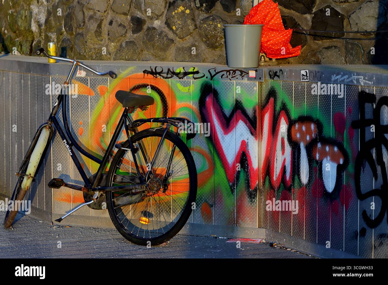 Altstadtrad lehnt sich an eine Graffiti-bedeckte Metallwand neben einem Damm, Amsterdam, Niederlande, warmes Abendlicht mit sichtbarem Eimer. Stockfoto