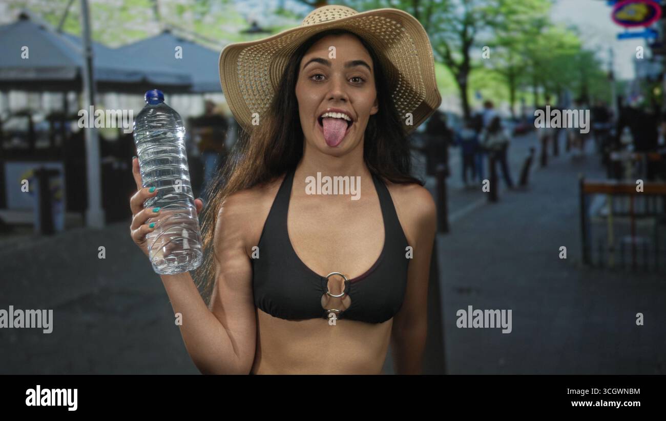 Die Frau hält die Wasserflasche in der erhobenen Hand mit der Zunge nach oben, während sie auf der Stadtstraße einen Sonnenhut trägt; Freude. Stockfoto