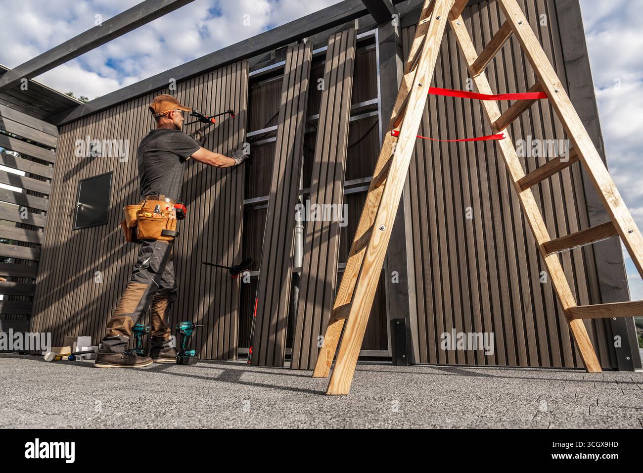 Ein gelernter Zimmermann arbeitet an der Installation von Holzpaneelen auf einer Außenkonstruktion unter blauem Himmel. Die Werkzeuge sind in der Nähe angeordnet, während er sorgfältig an misst Stockfoto