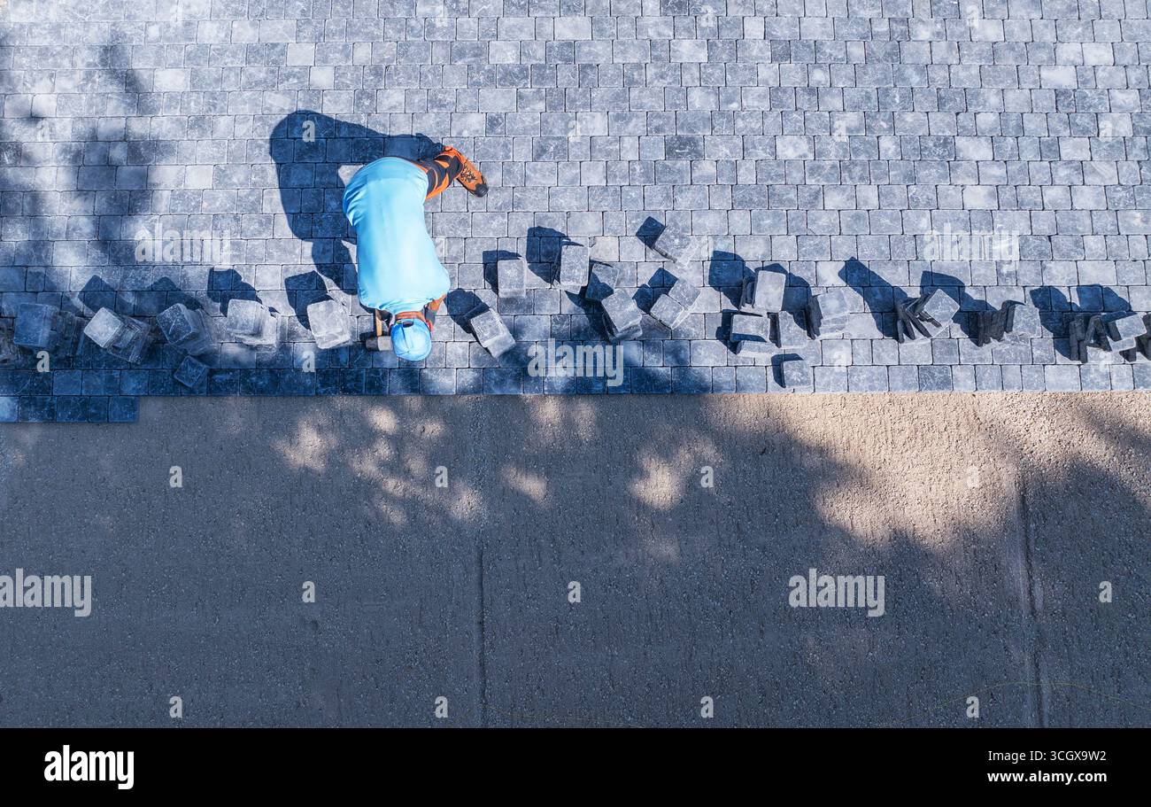 Ein Arbeiter legt Steinpflaster vorsichtig auf einen Weg, um Präzision und Ausrichtung unter der warmen Sonne zu gewährleisten. Schatten von Bäumen verleihen der Szene Tiefe. Stockfoto