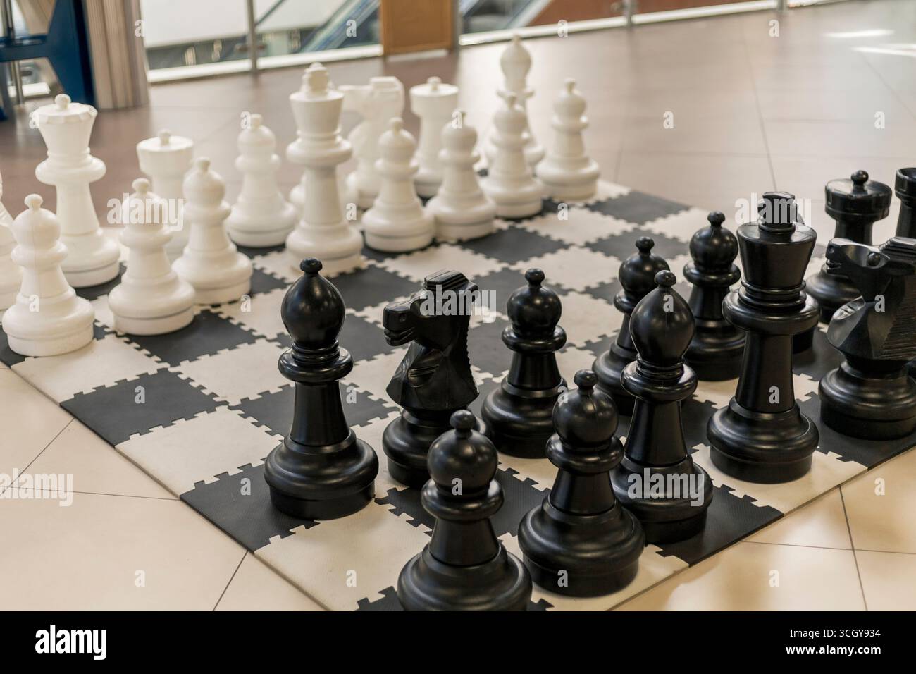 Ein großes Schachbrett mit übergroßen schwarzen und weißen Teilen ist drinnen ausgestellt. Die Spieler bereiten sich auf ein durchdachtes Spiel vor. Tageslicht Stockfoto