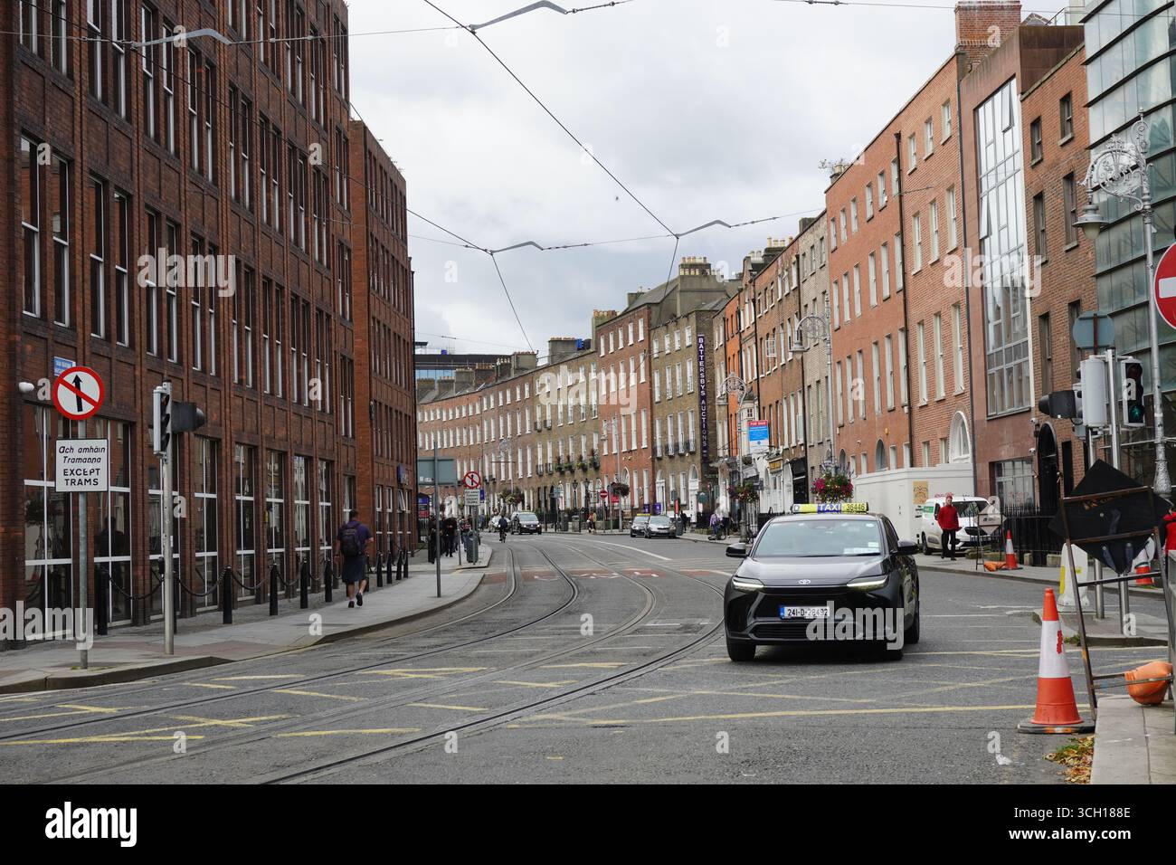 Eine Straßenszene in Dublin, in der ein schwarzes Taxi eine kurvenreiche Straße entlang fährt, die von klassischen georgianischen Backsteinhäusern und Straßenbahnleitungen gesäumt ist. Stockfoto