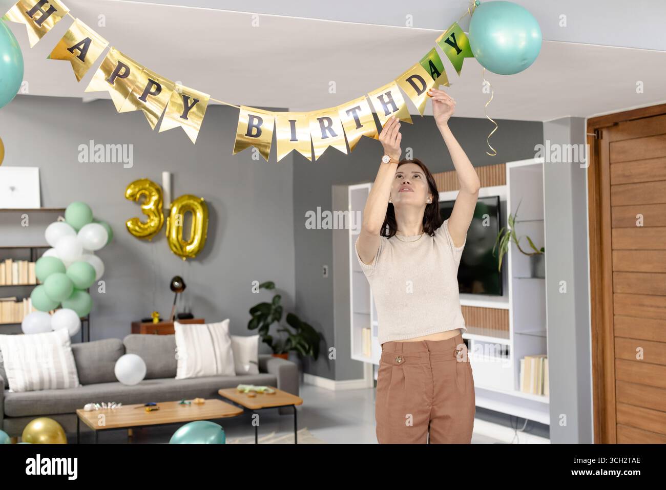 Frau hängte HAPPY BIRTHDAY Banner im Wohnzimmer mit goldenem 30 Ballon und mintgrünen Ballons Stockfoto