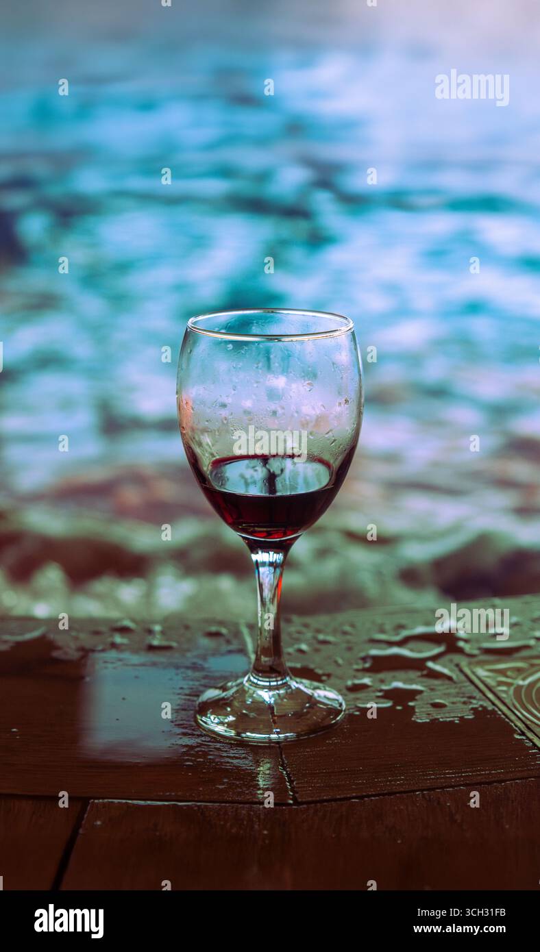 Ein Glas Rotwein auf einem nassen Holztisch mit blauem Wasser im Hintergrund eines beheizten Whirlpools. Stockfoto