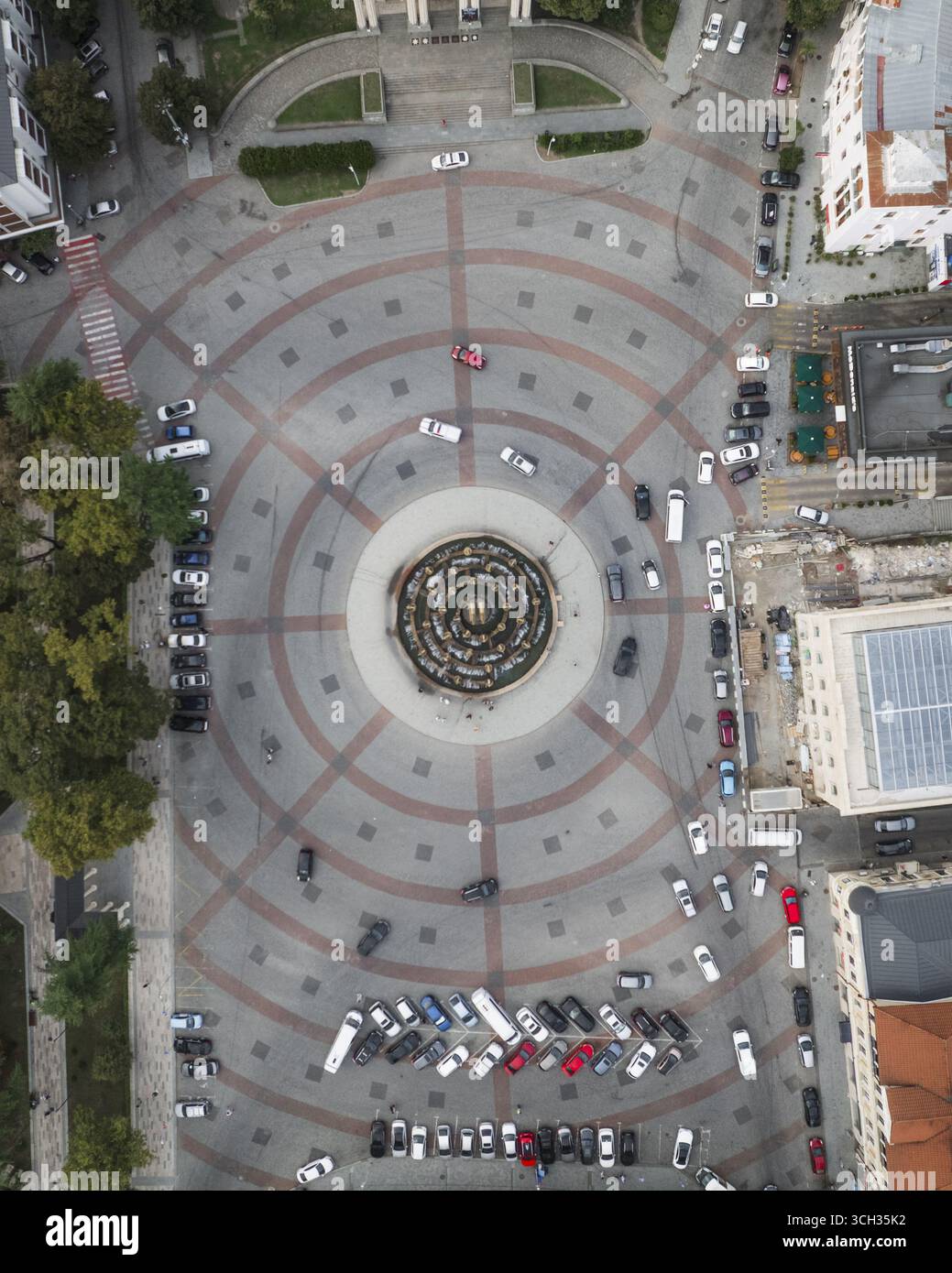 Aus der Vogelperspektive auf einen pulsierenden Kreisplatz voller Autos rund um den Colchis-Brunnen, eingerahmt von Gebäuden und grünen Bäumen, Kutaisi, Imereti, Georgia. Stockfoto