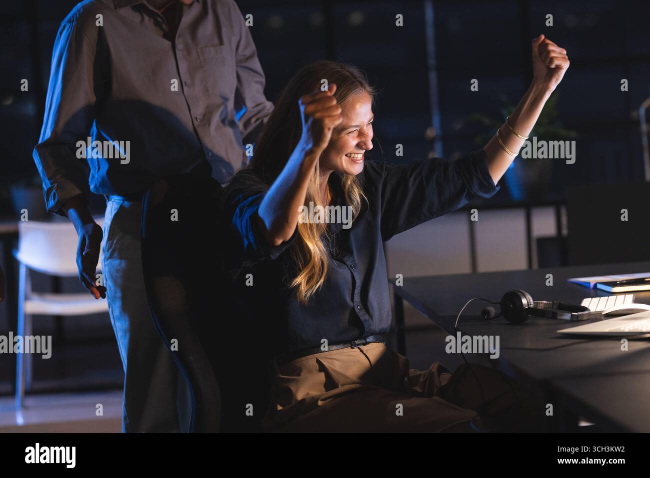 Die Frau feiert den Erfolg und erhebt triumphierend die Arme am Schreibtisch in der Nacht Stockfoto