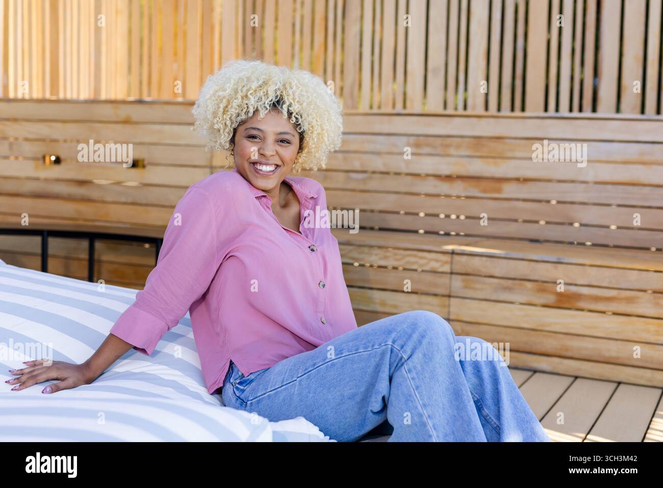 Lächelnde Frau in lässigem Outfit, die sich auf der Gartenbank im Freien entspannt und den Sonnenschein genießt Stockfoto
