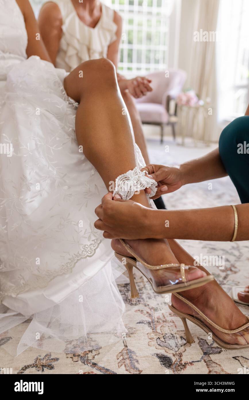 Braut, die Strumpfbänder von einem Freund angepasst und sich auf die Hochzeitszeremonie im Haus vorbereitet Stockfoto
