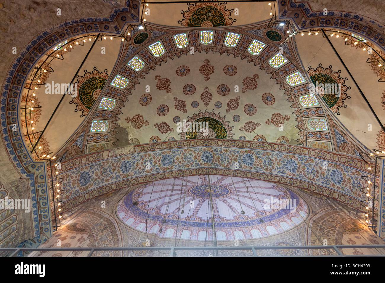 Türkei - Istanbul - Sultan-Ahmed-Moschee - bemalte Halbkuppel mit Arabesken, Fenstern und kalligraphischem Band, das sanft unter Lampen leuchtet Stockfoto
