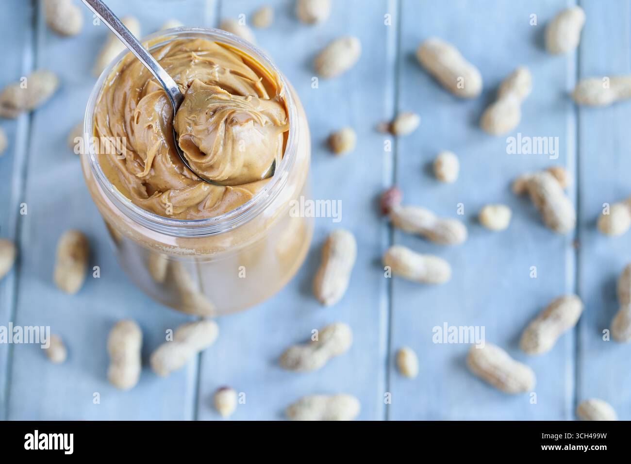 Erdnussbutter wird mit einem Löffel aus einem Plastikbehälter über einem blauen Tisch mit verstreuten Nüssen in der Schale aufgeschlüsselt. Selektiver Fokus mit Unschärfe Stockfoto