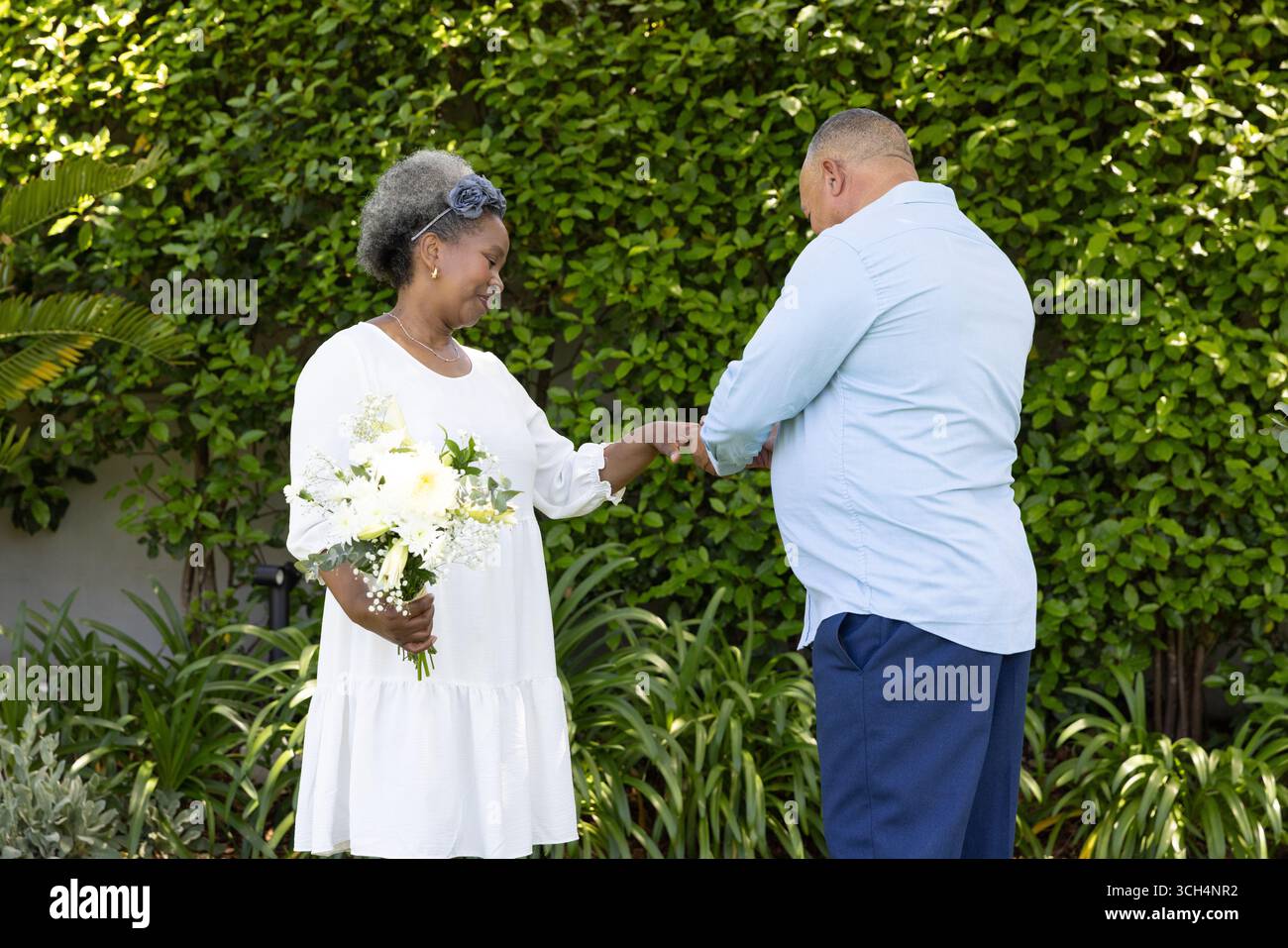 Seniorenpaar im Garten tauscht Ringe aus, Frau hält Blumenstrauß, beide lächeln warm Stockfoto