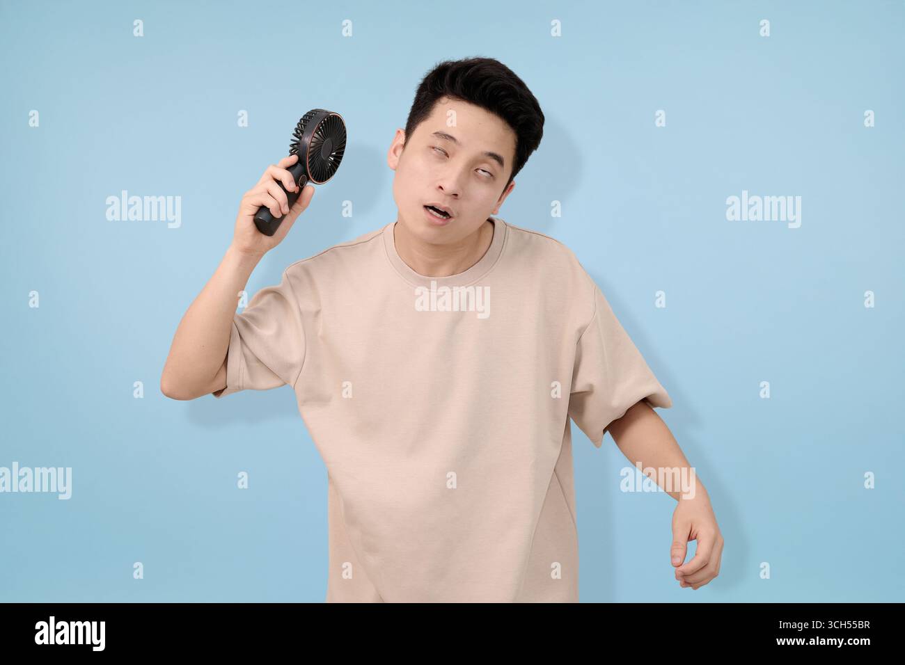 Ein junger Mann, der einen tragbaren Handventilator hält, der Erleichterung vor blauem Hintergrund ausdrückt Stockfoto