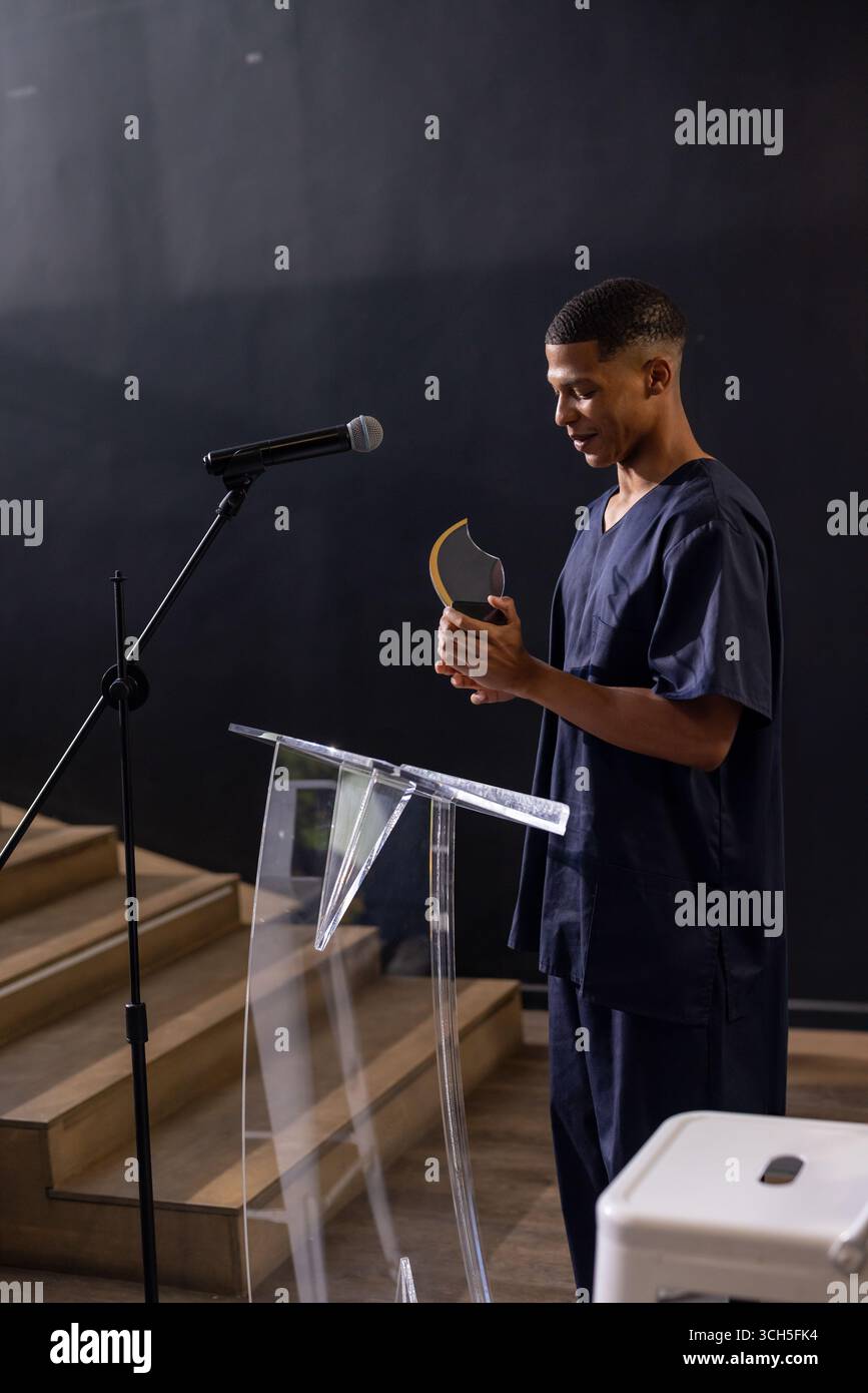 Mann, der auf der Bühne steht, auf dem klaren Podium mit Mikrofon, Gold-Schwarz-Preisträgertrophäe hält, Kopierraum Stockfoto