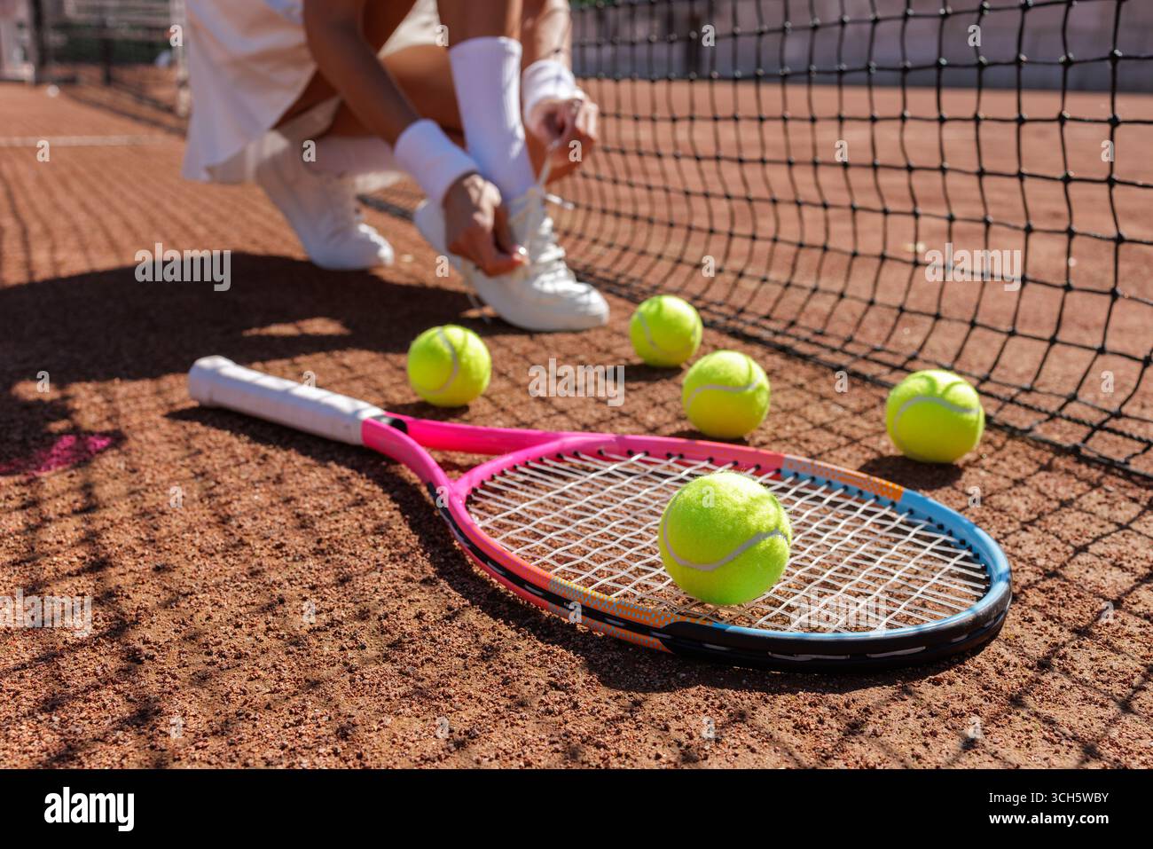 Schläger und bunte Bälle am Netz, während ein Spieler Schuhe auf Lehm legt, perfekt für Clubunterricht Coaching-Anzeigen und Turnieraktionen Stockfoto