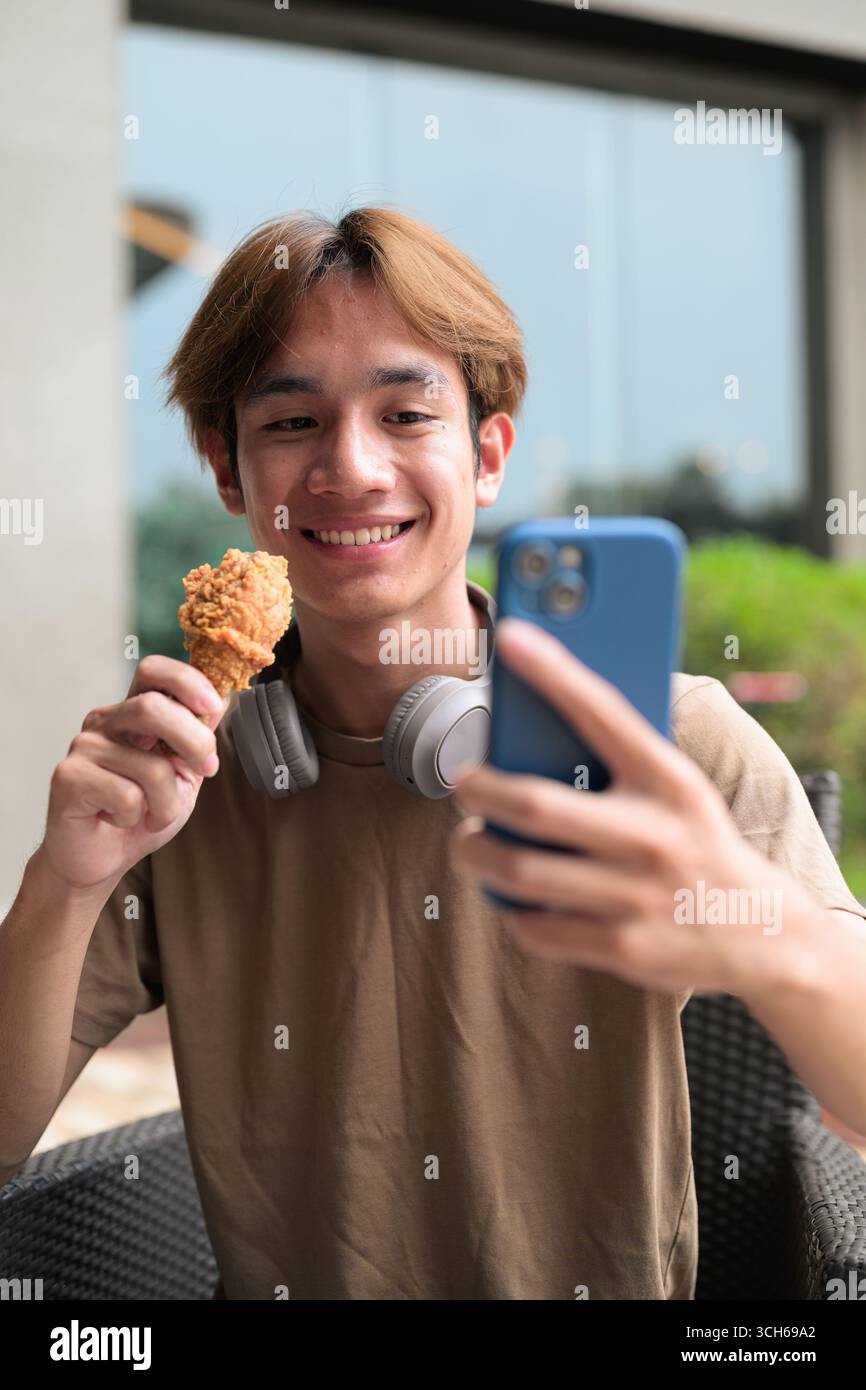Junger asiatischer Student Mann in braunem T-Shirt mit Kopfhörern, bereit, frittiertes Hähnchenkegel an einem Tisch im Freien für Fast Food, Snack und Freizeit Mea zu essen Stockfoto