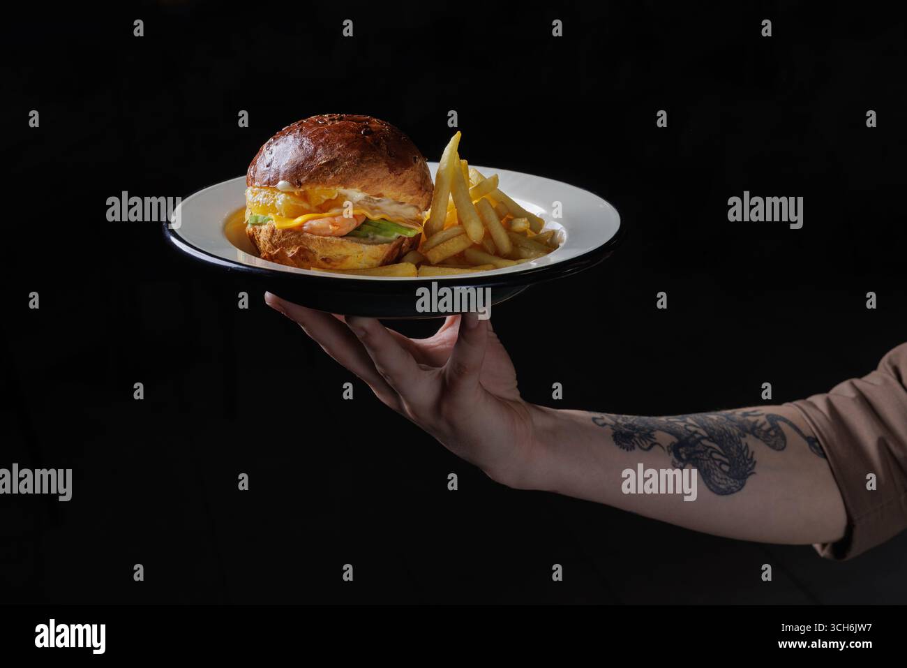 Gourmet-Burger mit Garnelen und Pommes auf Teller, serviert von tätowierter Hand auf dunklem Hintergrund mit künstlerischer Essenspräsentation. Stockfoto