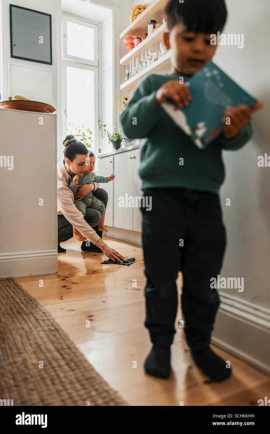 Frau, die den Jungen hält, während sie zu Hause den Boden der Küche reinigt Stockfoto