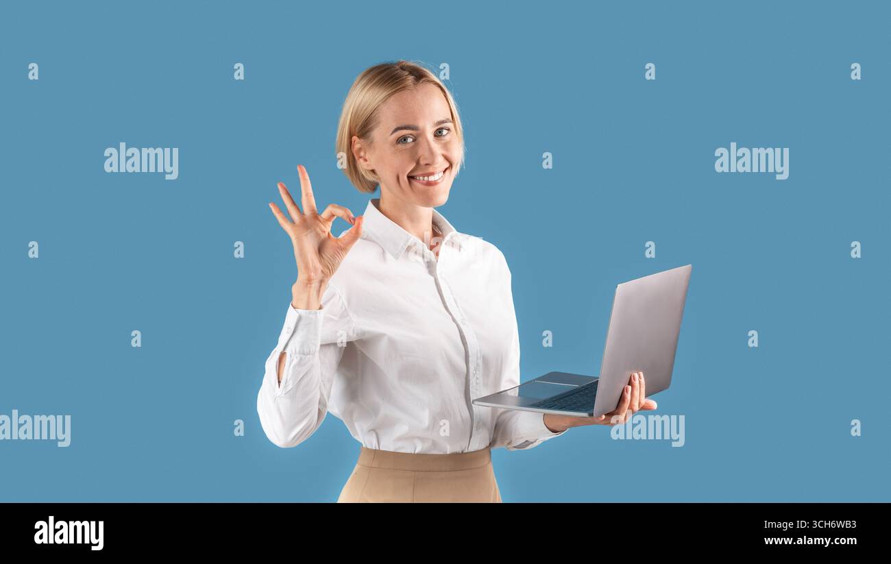 Fröhliche blonde Geschäftsfrau in formelle Kleidung zeigt OK Geste während Arbeiten auf Laptop über blauem Studio Hintergrund Stockfoto