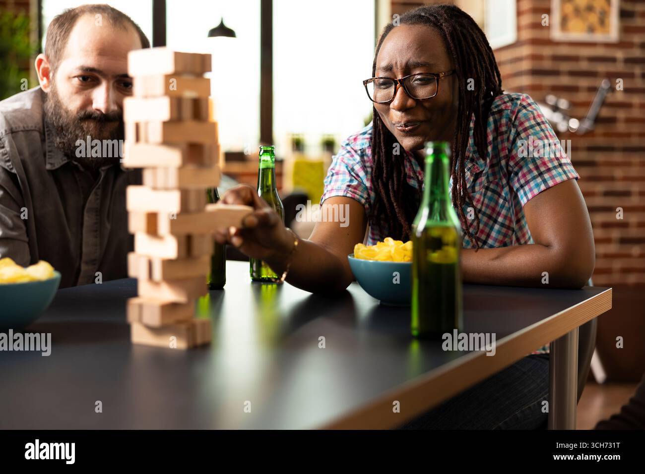 Eine afroamerikanische Frau mit nervösen Ausdrücken zieht Holzblock aus dem Jenga-Turm, während ihr kaukasischer Freund beobachtet. Junge schwarze Dame mit Brille, konzentriert und angespannt während des Tischspiels. Stockfoto