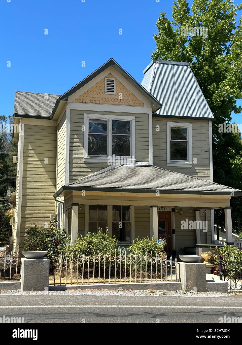 Ein historisches zweistöckiges Holzhaus mit Dach im Turmstil, überdachter Veranda und dekorativer Dekoration, fotografiert an einem sonnigen Tag mit blauem Himmel. - Smartphone-aufgenommenes Stockfoto