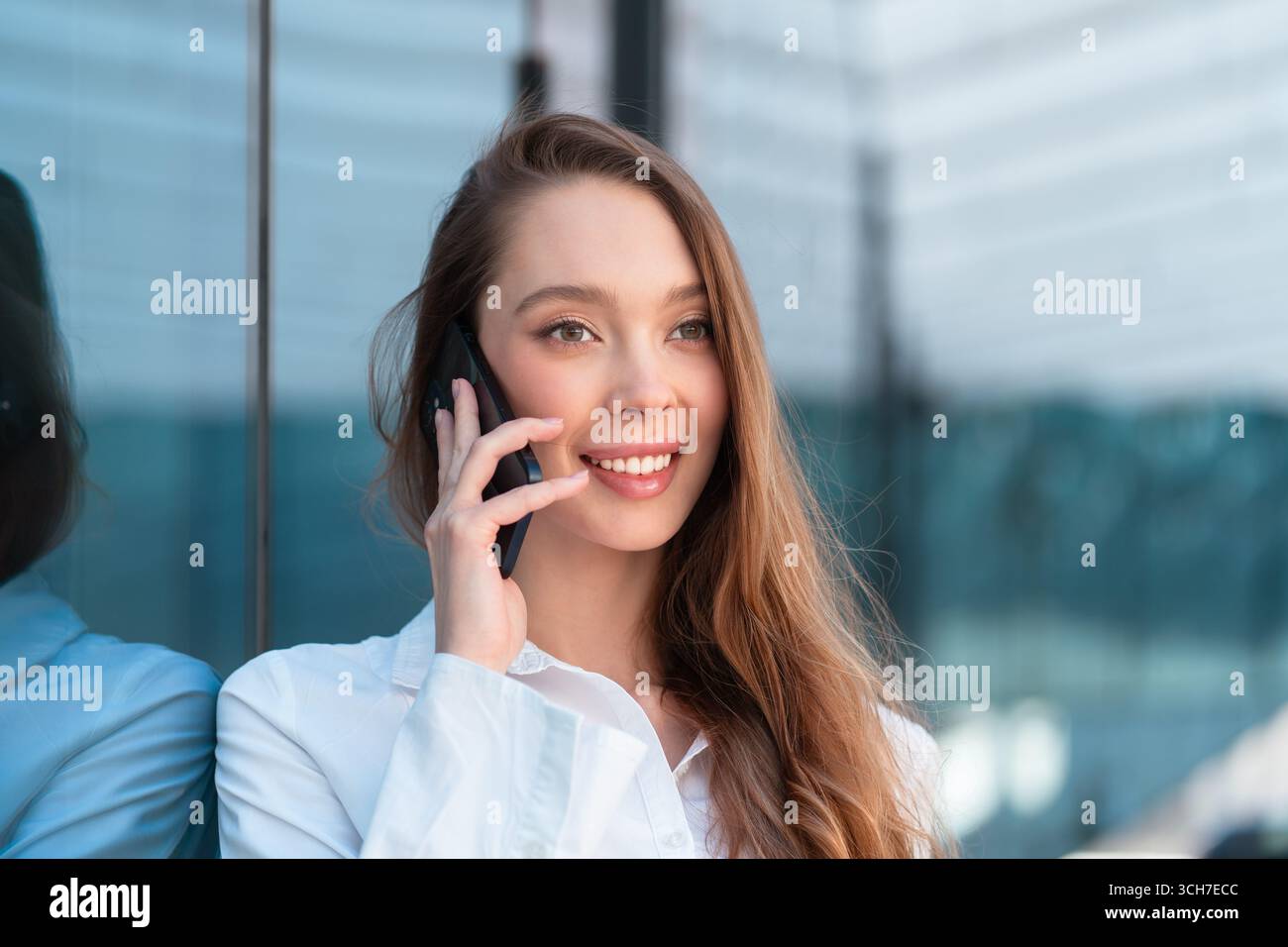 Glückliche Geschäftsfrau in weißem Hemd in der Nähe des Bürogebäudes, die ihr Smartphone hält und während des Telefongesprächs lächelt, was Selbstvertrauen und Verbindung zeigt Stockfoto