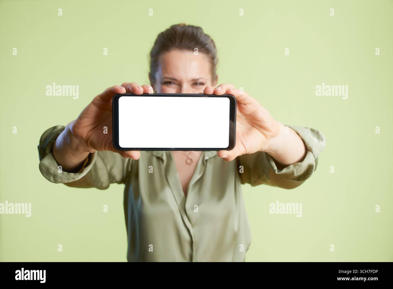 Eine Frau hält ein Smartphone horizontal, um seinen leeren Bildschirm zu zeigen. Informative Technologie-Anzeige vor einem sanften grünen Hintergrund. Stockfoto