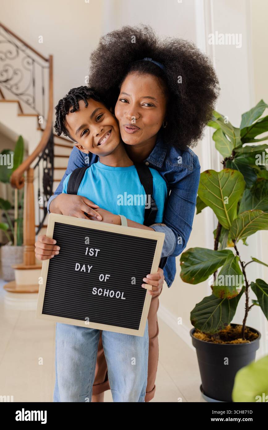 Afroamerikaner Mutter umarmt den Sohn mit dem 1. Schultag Schild, beide lächeln Stockfoto