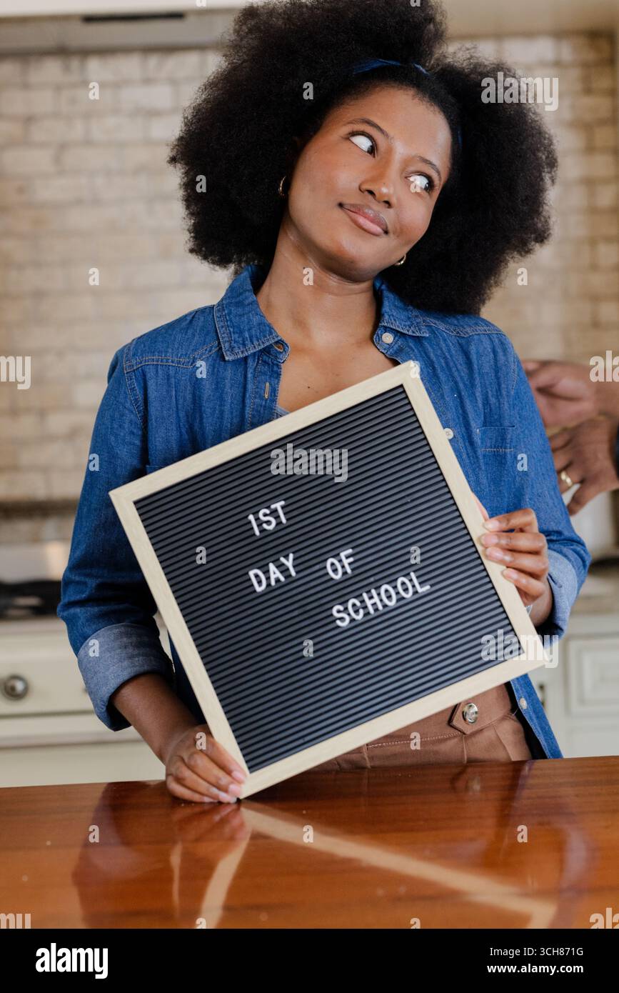 Afroamerikanerin, die ein Schild für den 1. Schultag hält, sieht in der Küche nachdenklich aus Stockfoto