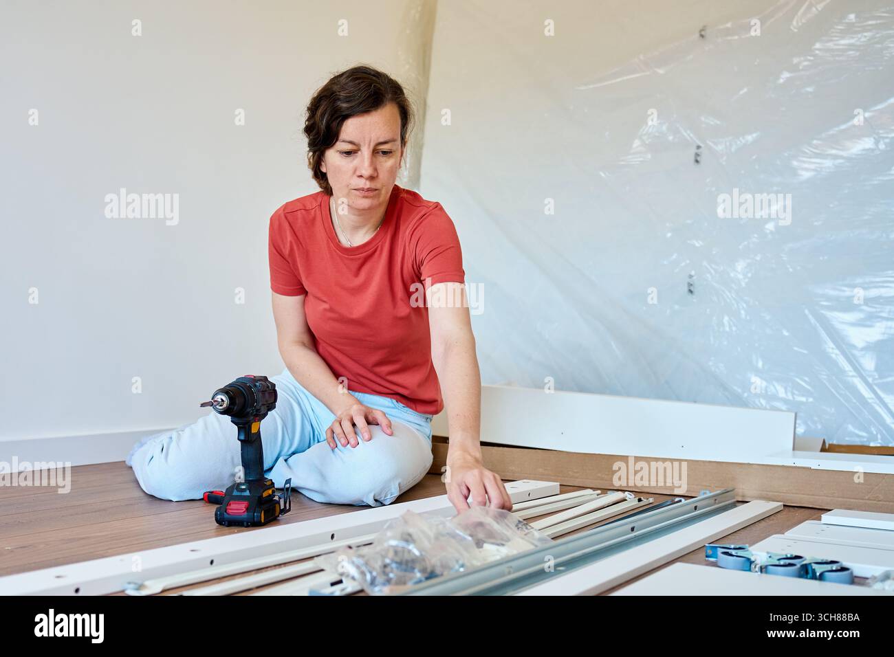 Frau, die Teile für die Möbelmontage zu Hause auspackt. Frau, die auf dem Boden sitzt und Bettteile, Räder und Befestigungsteile hält. Das Konzept des Heimwerkerimprovisierens Stockfoto