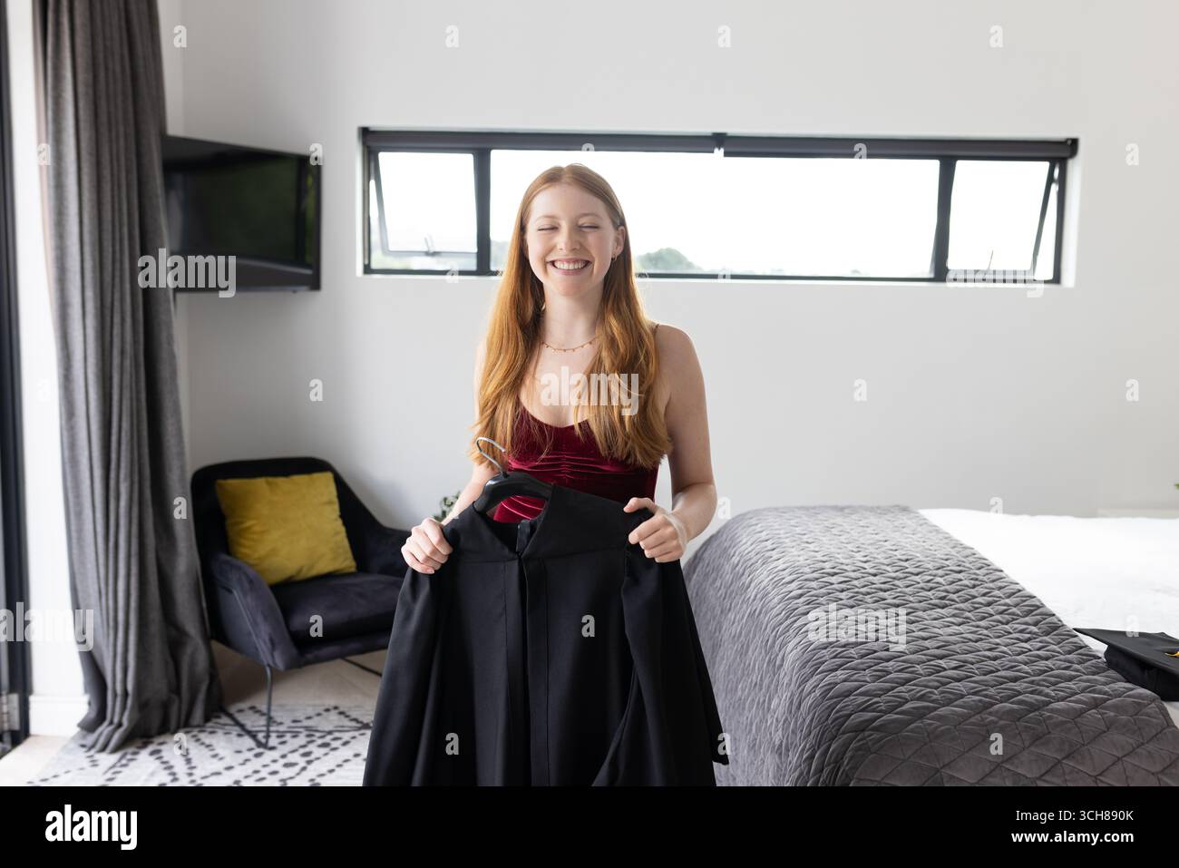 Teenager im Schlafzimmer lächelt, während sie Kleid hält und sich auf die Feier vorbereitet Stockfoto