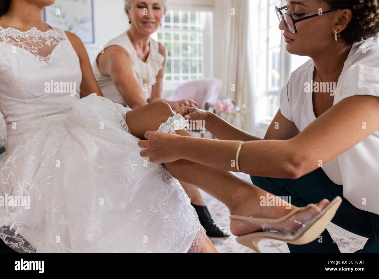 Braut trägt Spitzengrüpp, während Freunde helfen, sich auf die Hochzeitszeremonie vorzubereiten Stockfoto